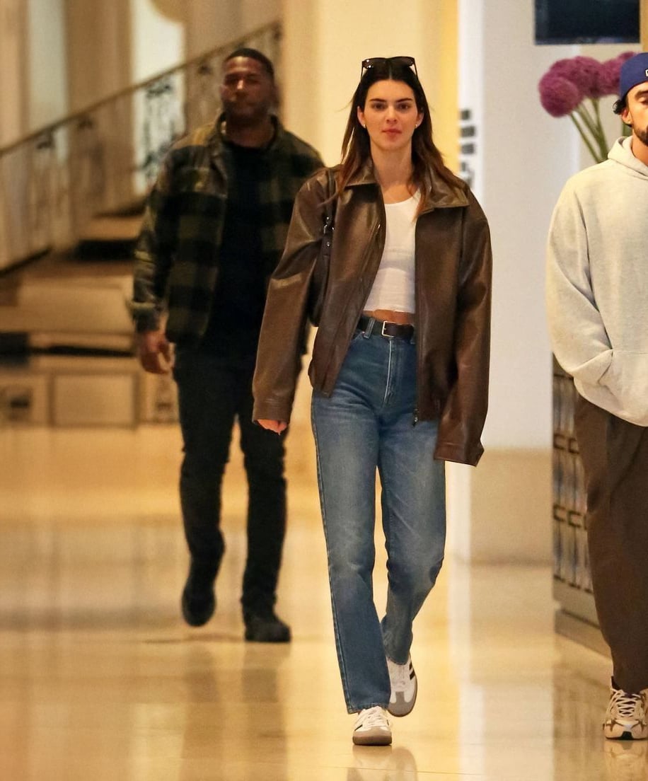 A young woman wearing a brown leather jacket, white top, blue jeans, and white sneakers walks indoors, with sunglasses on her head. A man in a plaid shirt and another person in a hoodie are visible in the background.