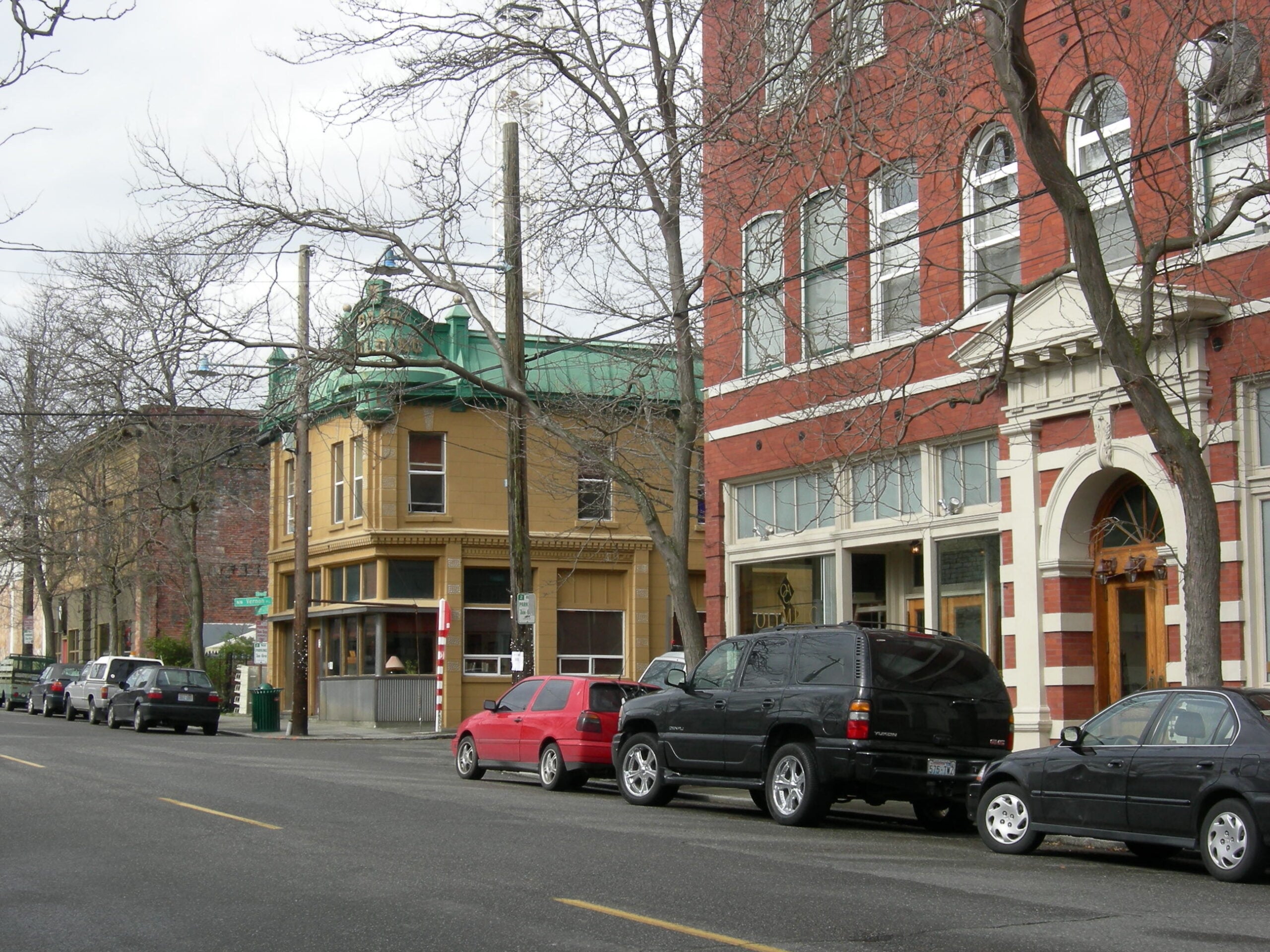 A city street lined with parked cars, leafless trees, and old brick buildings, including a red and white building and a yellow one with a green roof, under a cloudy sky.