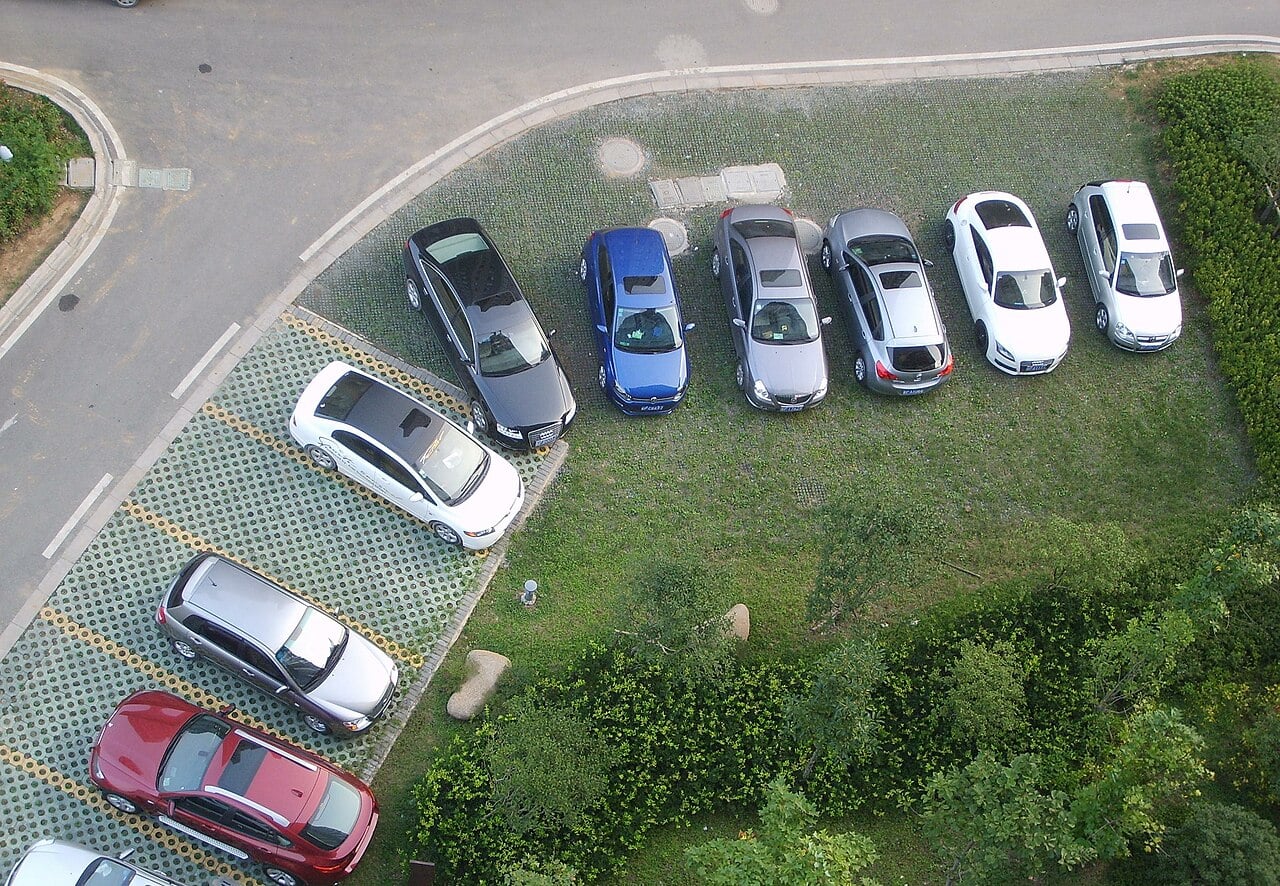 Aerial view of a parking lot with cars parked in organized rows; some cars are on a paved surface while others are parked on a grassy area near green shrubs and trees.