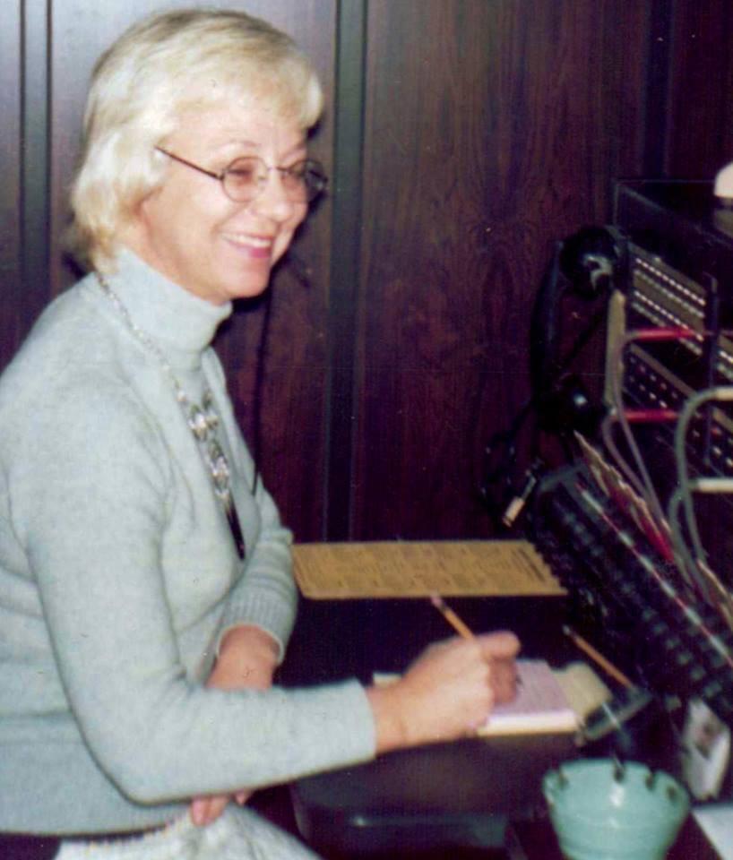 An older woman with short light hair and glasses sits smiling at a vintage telephone switchboard, writing on a notepad with a pencil, surrounded by cords and equipment.
