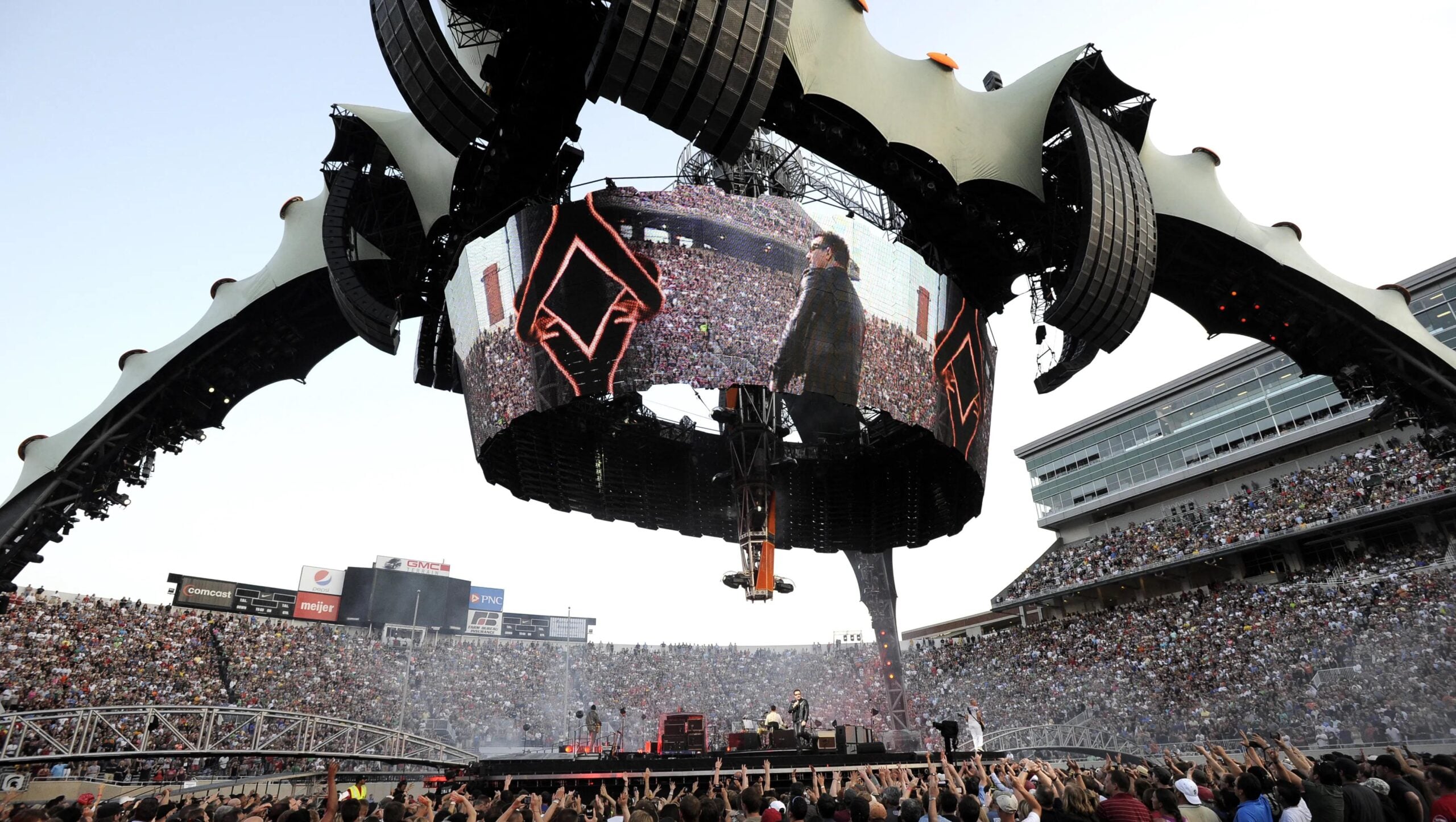 A massive stadium concert with a futuristic, claw-like stage structure and a large circular screen displaying a performer. The stage is surrounded by a cheering, packed crowd and stadium seating filled with fans.