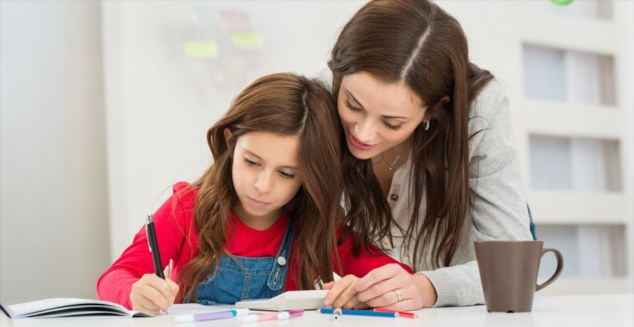 A woman helps a young girl with homework at a table. The girl writes in a notebook while the woman leans over, guiding her. Colored pens and a brown mug are on the table. Both appear focused and engaged.
