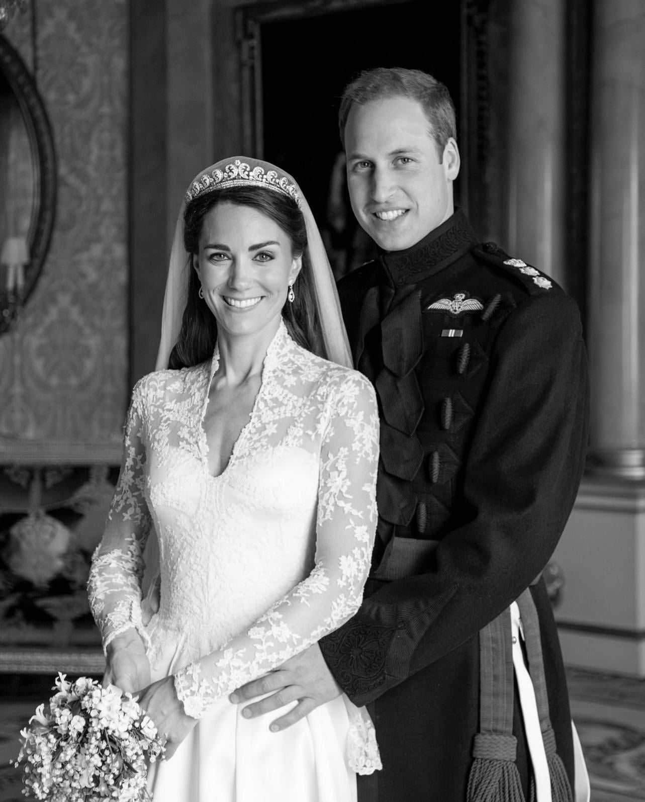 A bride in a lace wedding dress and tiara stands next to a groom in a military uniform. They are both smiling, posing indoors in an elegant, ornate room. The groom’s hand rests on the bride's waist.