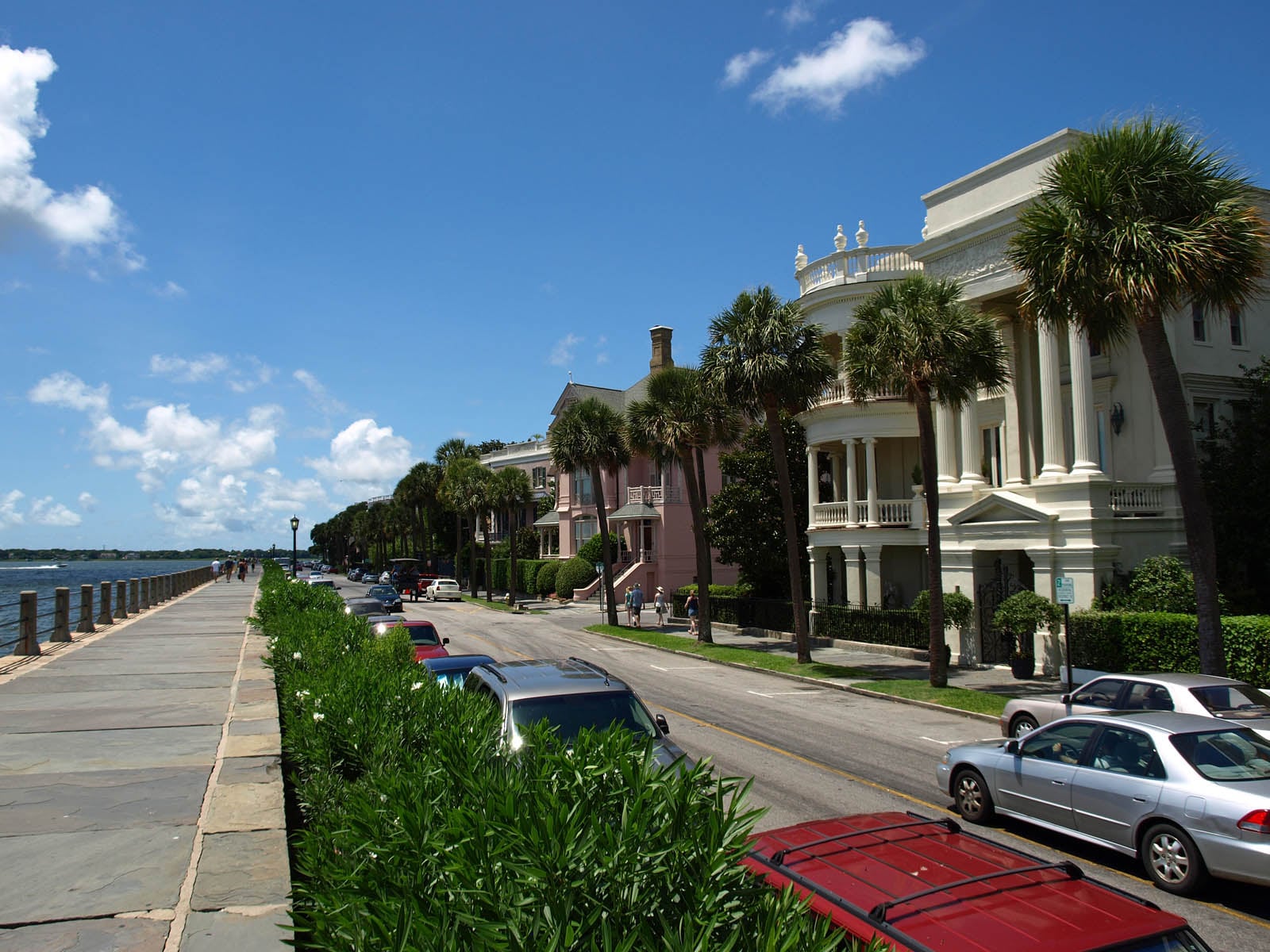 Historic mansions with columns and balconies line a palm tree-lined street beside a waterfront promenade under a bright blue sky with scattered clouds. Cars are parked along the road.