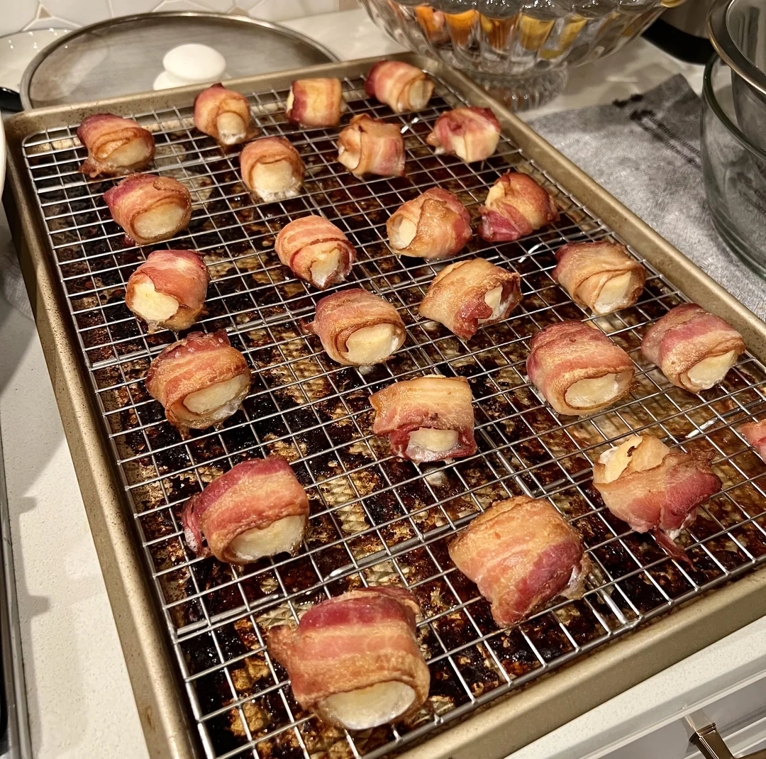 A wire rack on a baking sheet holds several bacon-wrapped cheese bites, freshly baked and golden brown. The baking sheet rests on a kitchen counter, with bowls and kitchenware in the background.
