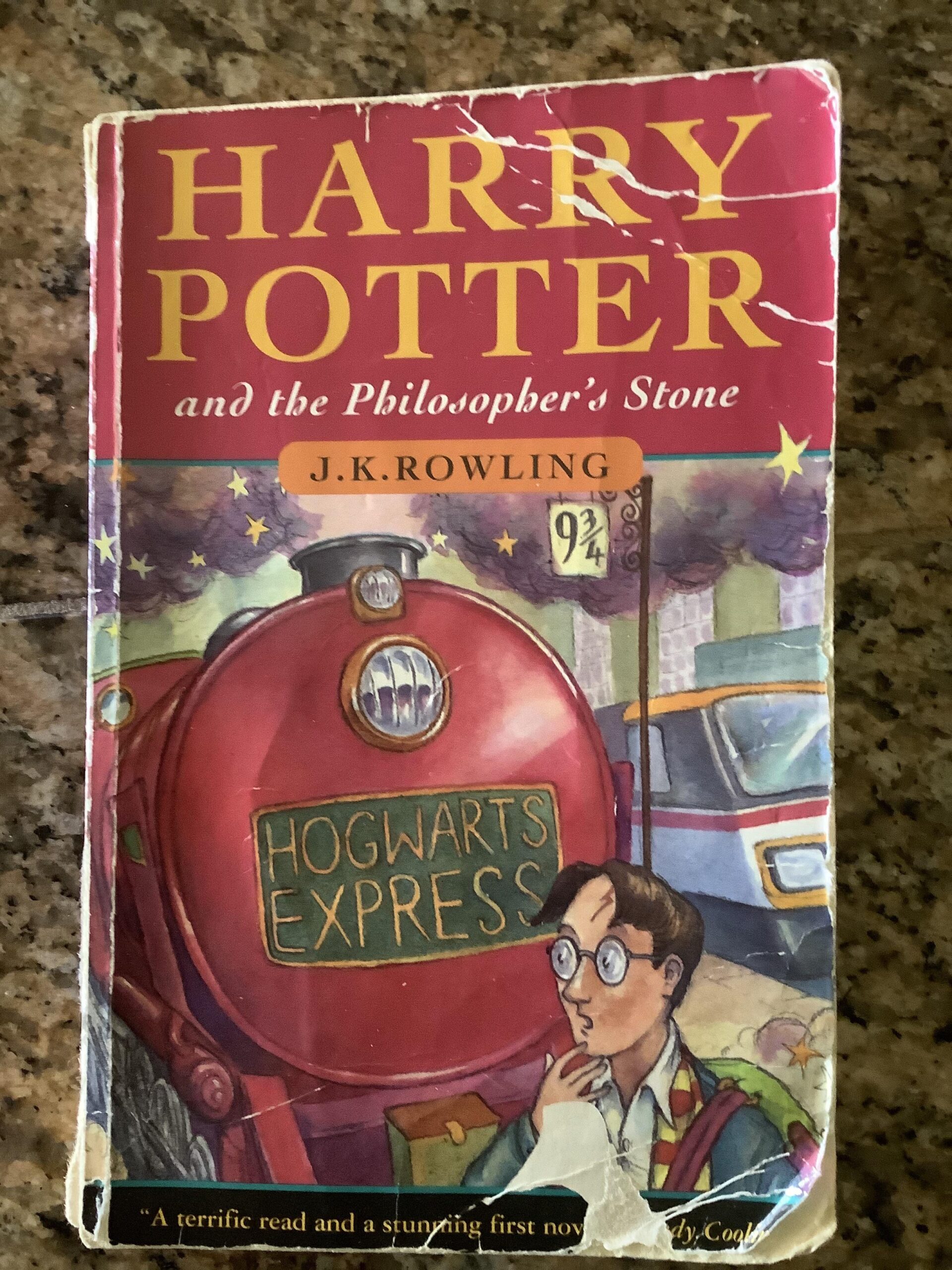 A worn copy of "Harry Potter and the Philosopher’s Stone" by J.K. Rowling lies on a granite surface. The cover shows Harry Potter at Platform 9¾ beside the red Hogwarts Express train.