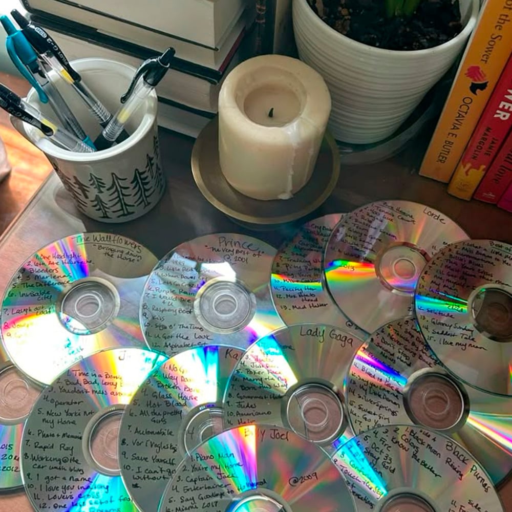 A collection of handwritten CDs with music playlists sits on a wooden table next to a candle, a cup holding pens, potted plants, and stacked books.