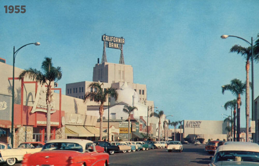 A 1955 street scene with classic cars parked along palm tree-lined storefronts. A tall building with a "California Bank" sign stands in the background under a clear blue sky.