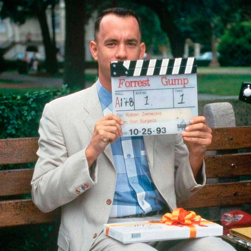 A man in a light suit sits on a bench holding a movie clapperboard labeled "Forrest Gump." A box of chocolates with a red ribbon rests on his lap. Trees and grass are visible in the background.