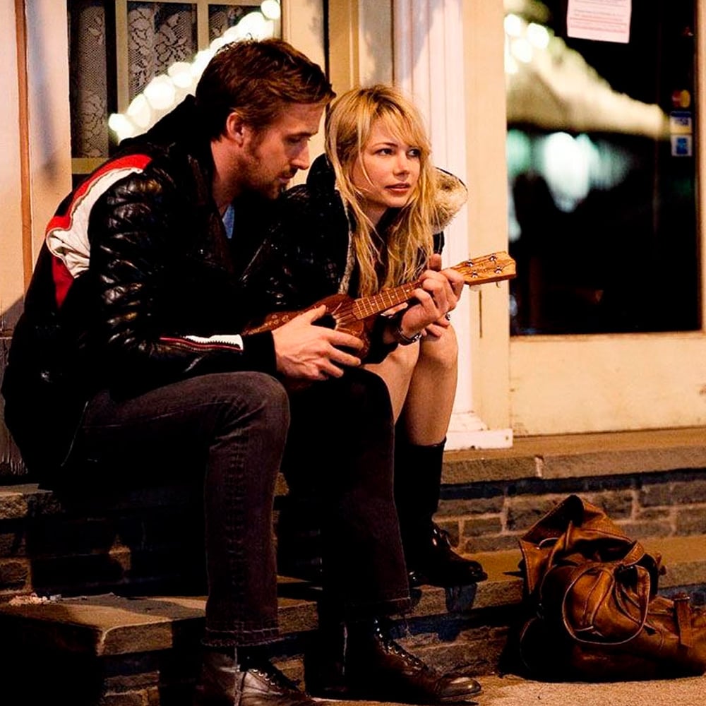 A man and a woman sit together on outdoor steps at night. The man plays a small guitar while the woman watches and smiles slightly. A brown bag rests on the ground beside them. Warm lights glow in the background.