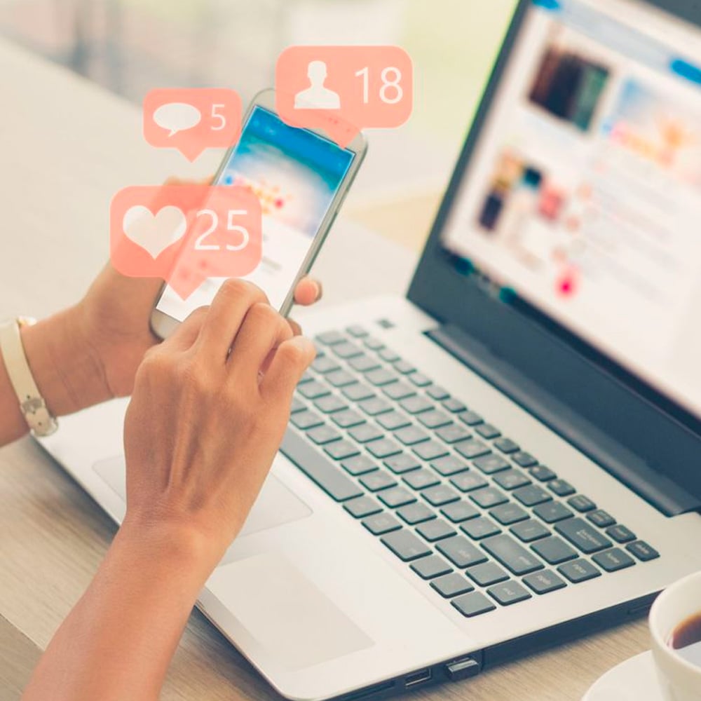 A person uses a smartphone near an open laptop. Social media notification icons for likes, comments, and followers appear above the phone screen, indicating active engagement.