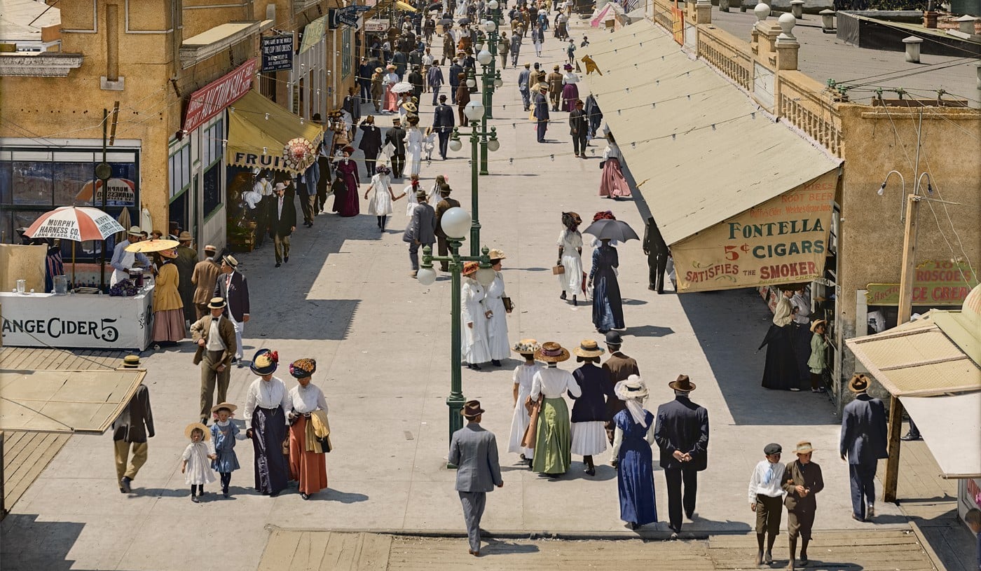A busy outdoor market scene from the early 1900s shows men, women, and children in period clothing walking and shopping among vendor stalls and shop signs on a wide pedestrian street.