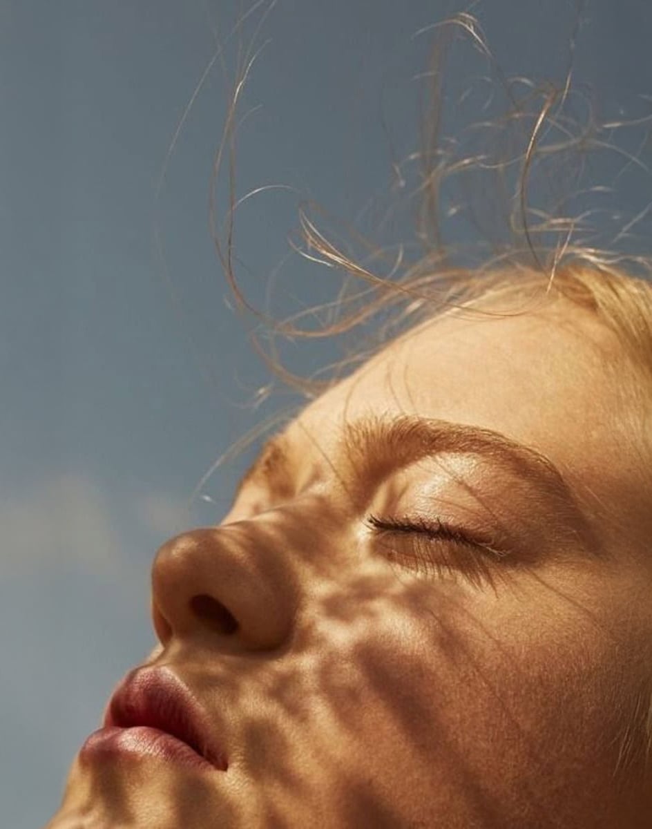 Close-up of a person's face with eyes closed, sunlight casting delicate shadow patterns on their skin, and wisps of hair blowing in the gentle breeze against a clear blue sky.