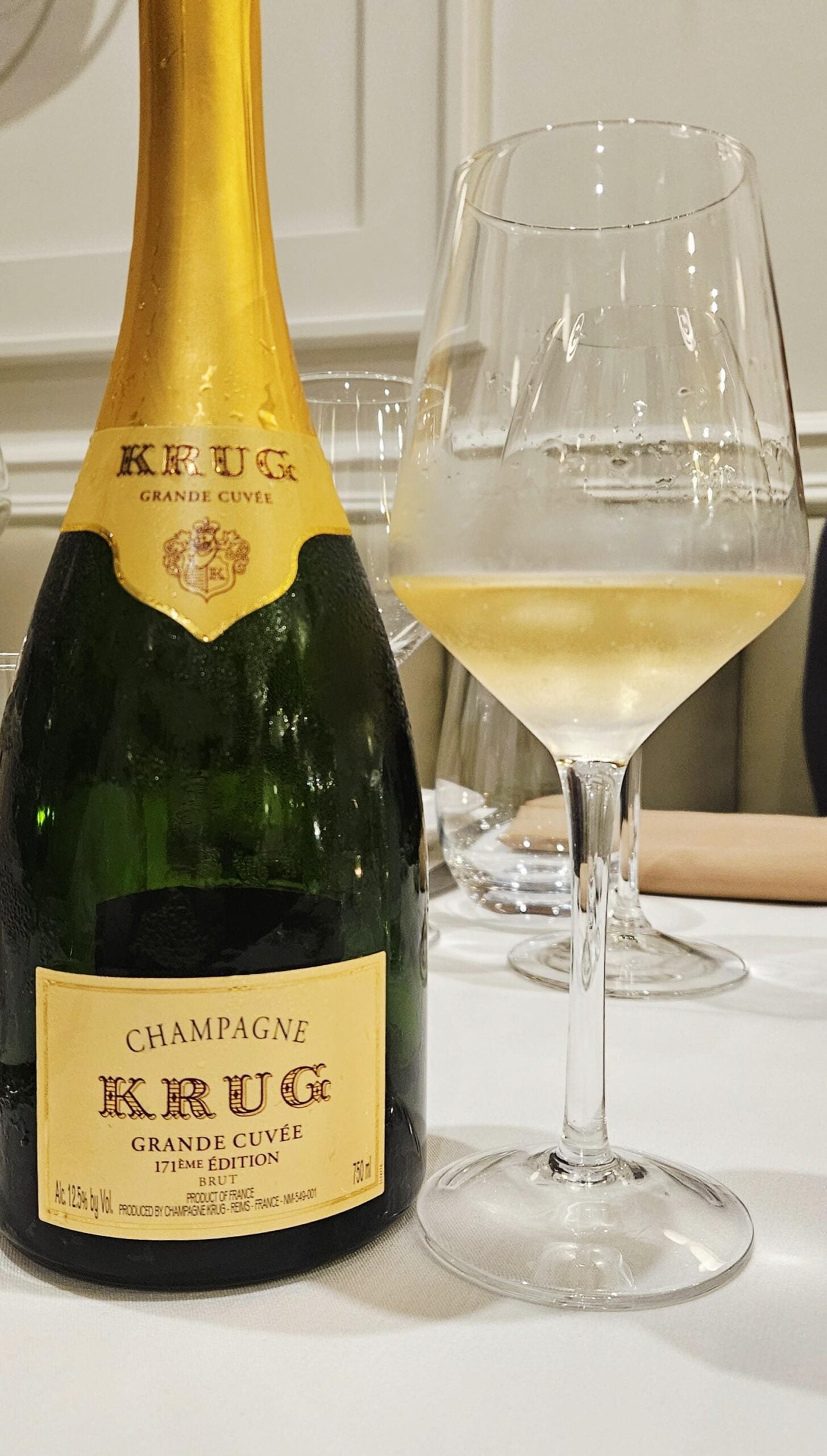 A bottle of Krug Grande Cuvée Champagne is placed on a white tablecloth next to a filled wine glass. The background shows more glassware and a neutral, elegant setting.