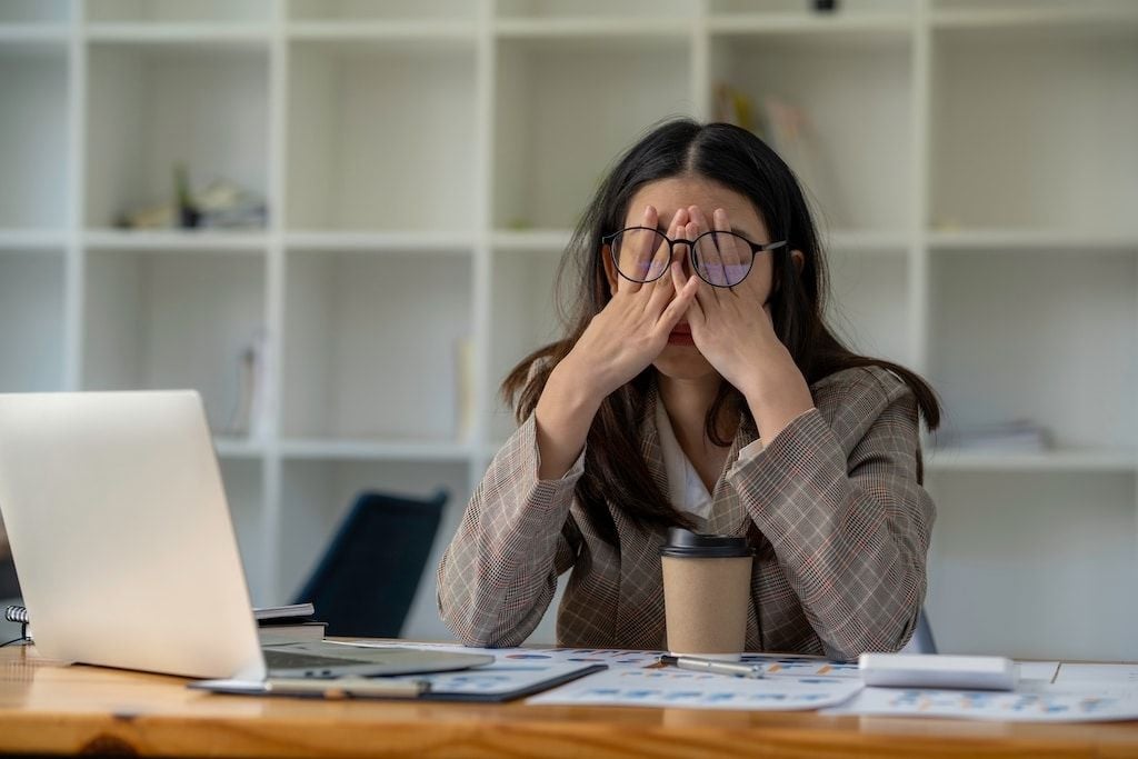 A woman wearing glasses and a plaid blazer sits at a desk with a laptop, rubbing her eyes in fatigue or frustration. A coffee cup, papers, and a pen are on the desk, with shelves in the background.
