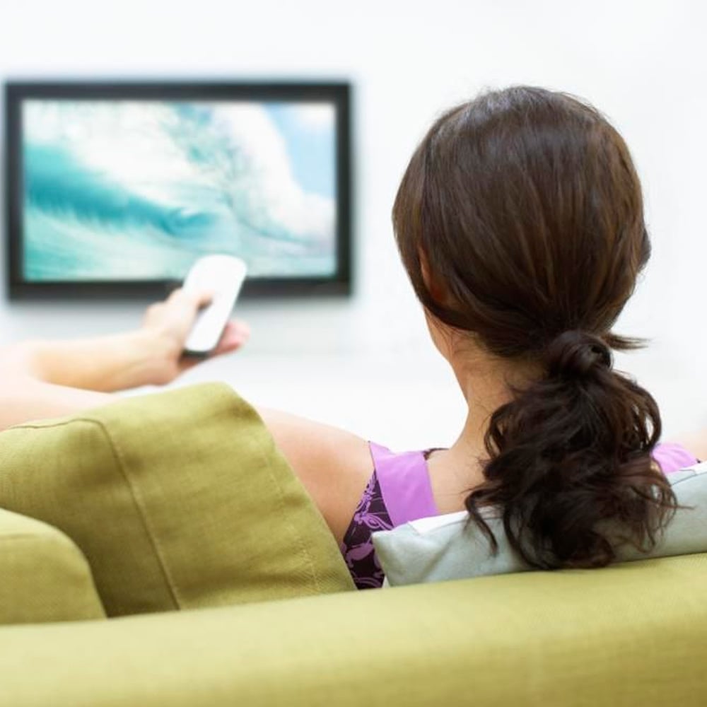 A woman with brown hair in a ponytail sits on a green sofa, holding a remote control and watching a TV screen showing an ocean wave.