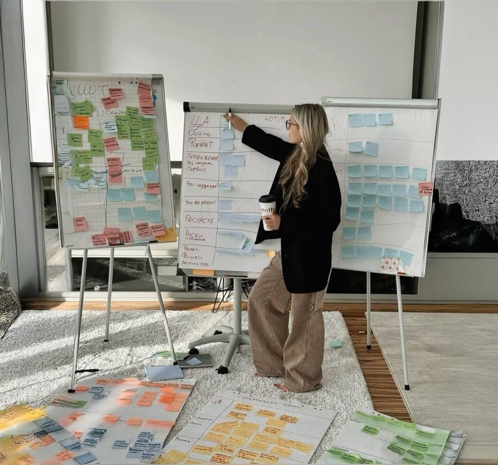 A person stands in front of whiteboards and flipcharts covered with colorful sticky notes and papers, holding a coffee cup and pointing to a note, appearing to lead a brainstorming or planning session.