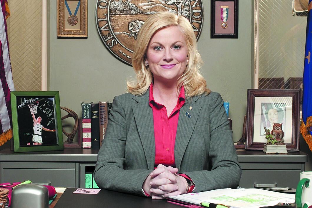 A woman with blonde hair in a gray blazer and red shirt sits at a desk in an office, hands clasped. Behind her are family photos, books, an American flag, and a large seal on the wall.