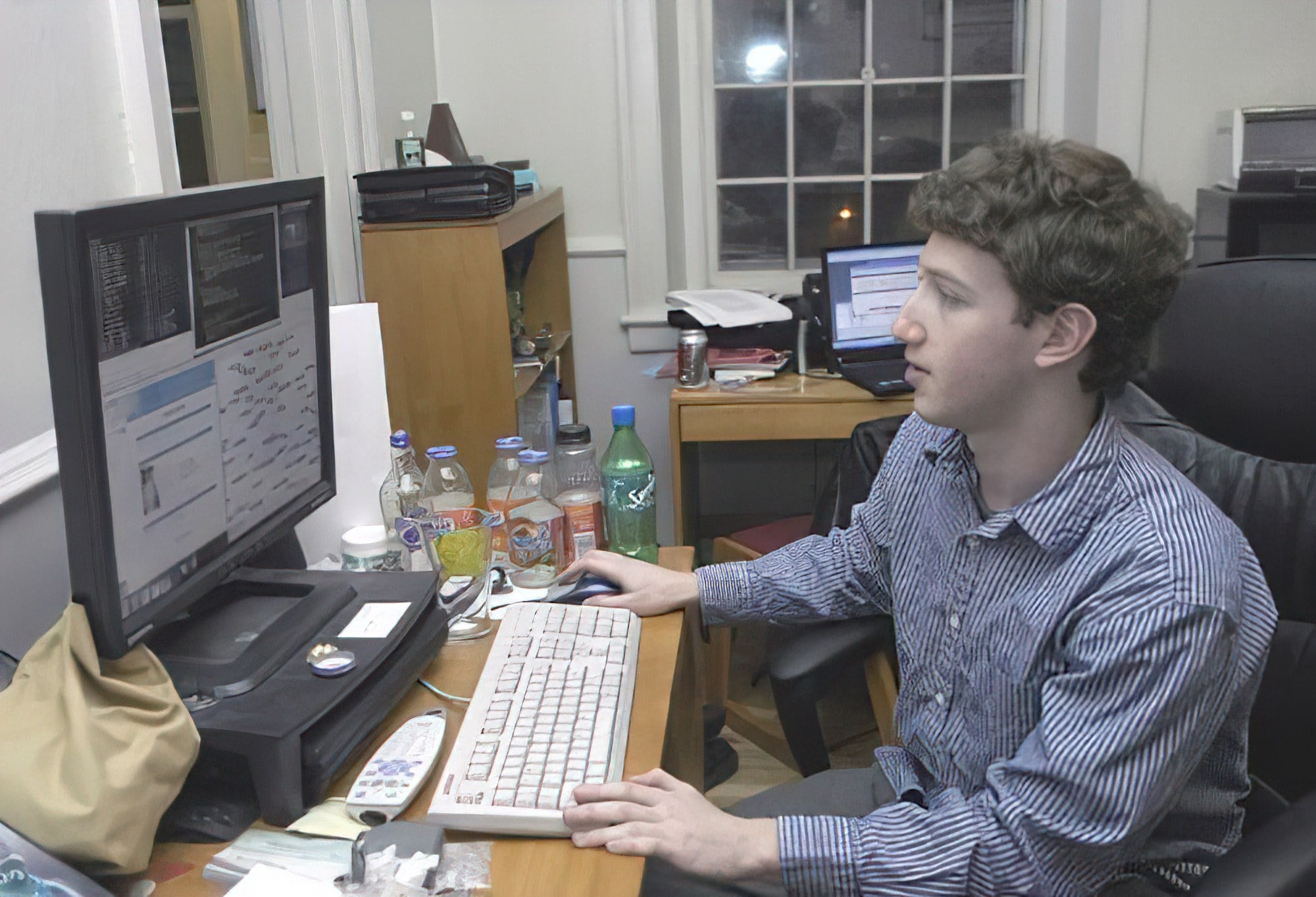 A young man sits at a desk using a computer with multiple screens. The desk is cluttered with bottles, papers, and electronic equipment. Another desk with a computer is in the background near a window.