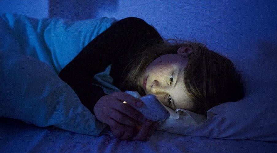A person lying in bed at night, illuminated by the blue glow of a phone screen, looks at their device while resting their head on a pillow.