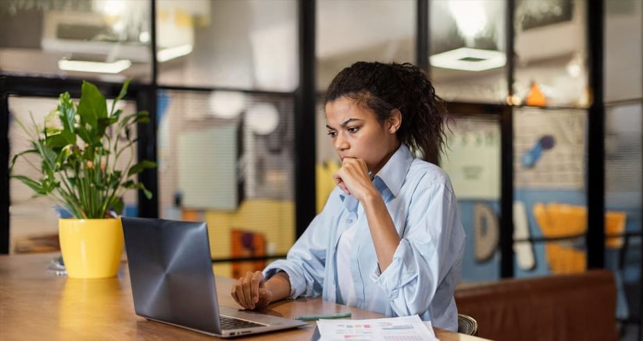 A woman sits at a desk in a modern office, looking thoughtfully at her laptop. Papers and a yellow potted plant are on the desk, and large glass windows are in the background.