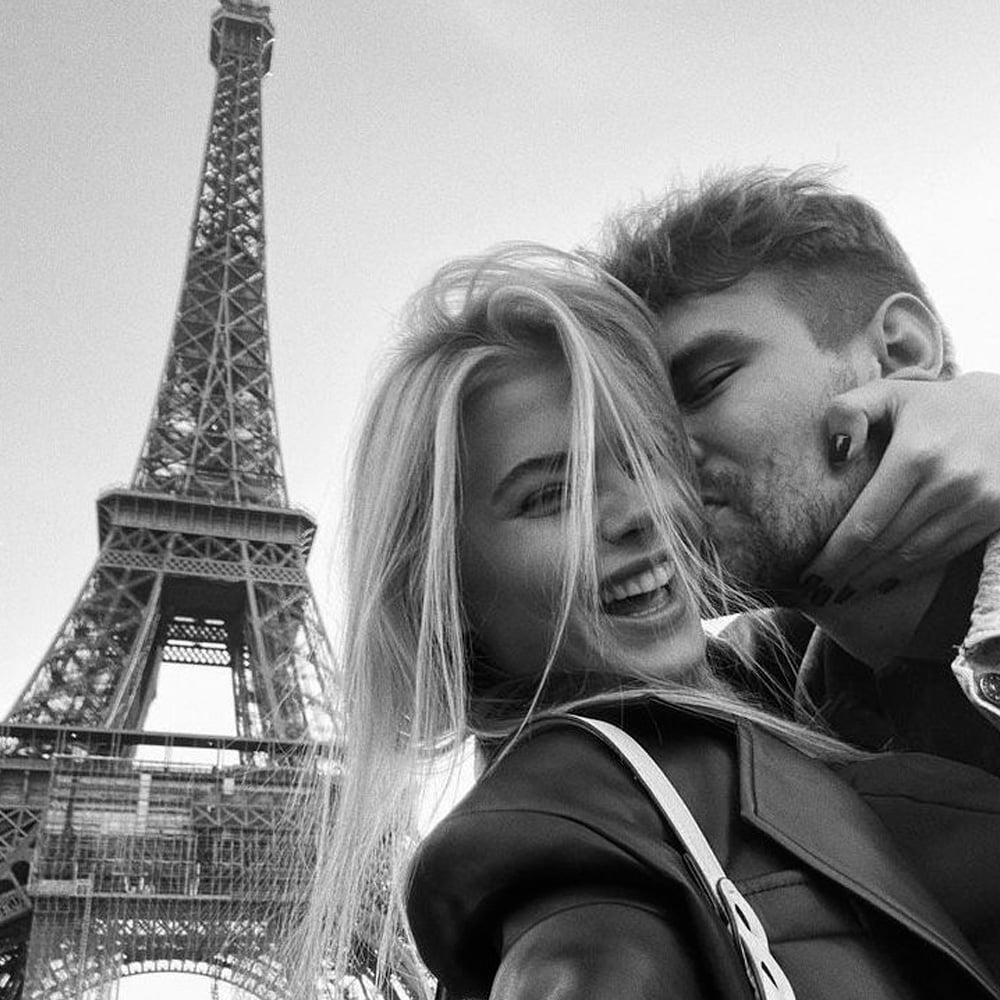 A couple takes a joyful selfie in front of the Eiffel Tower. The woman smiles at the camera while the man kisses her cheek. The photo is in black and white.