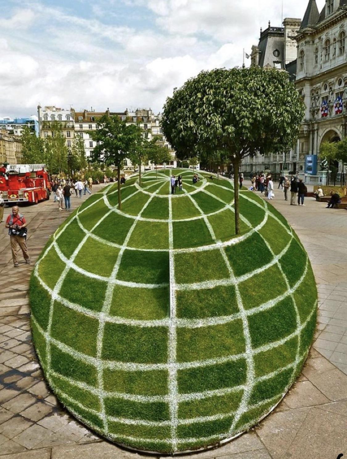 A grass-covered mound shaped like a globe with white grid lines stands in a city square, topped with trees and surrounded by people and historic buildings.