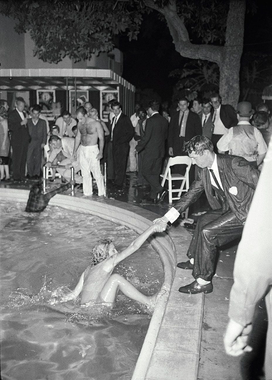 A man in a wet suit kneels by a pool, handing a drink to a woman in the water as a crowd of people in formal attire watch at night. Some people stand, others sit, and the setting appears to be a lively party.