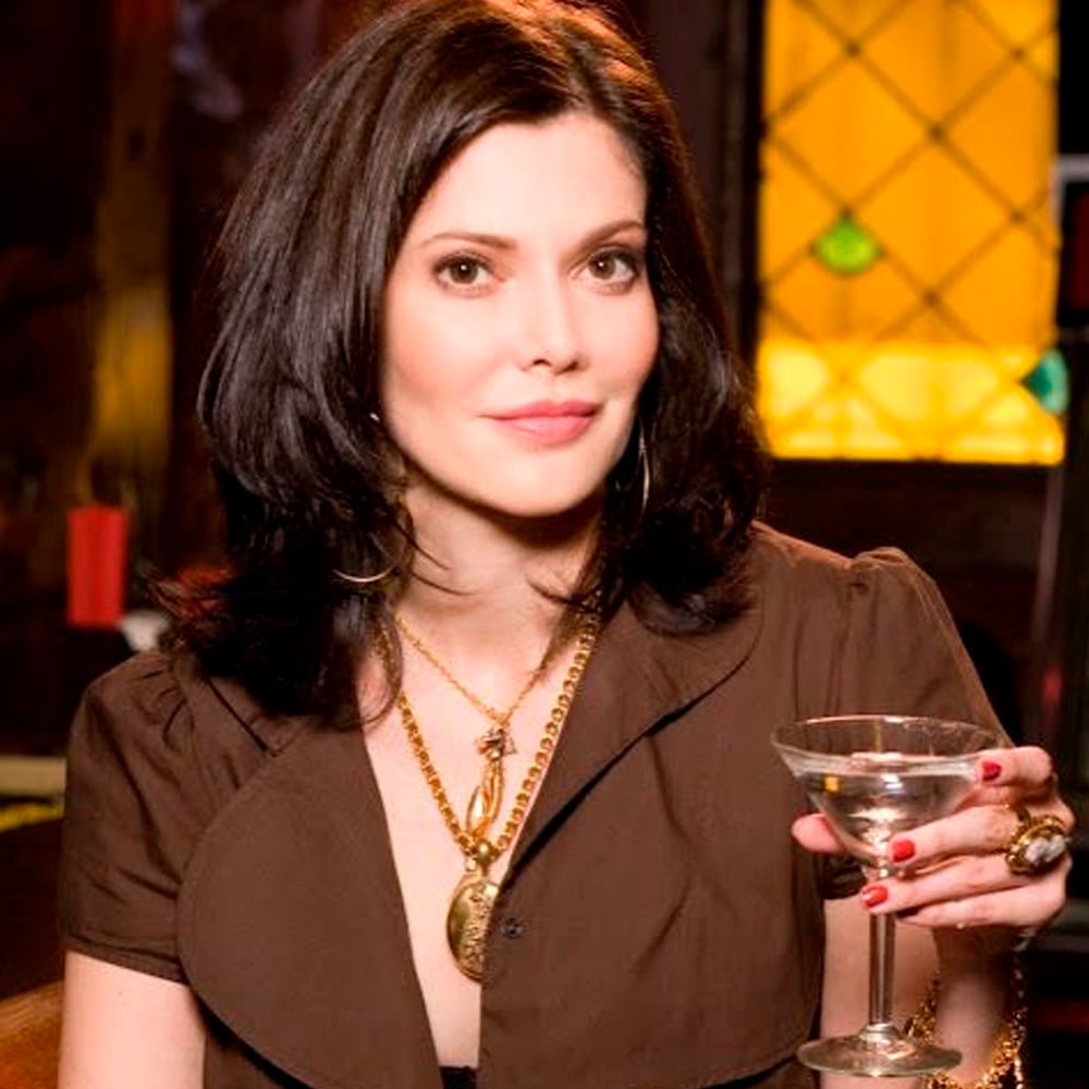 A woman with shoulder-length dark hair wearing a brown top and gold jewelry holds a martini glass, sitting indoors with dim, warm lighting in the background.