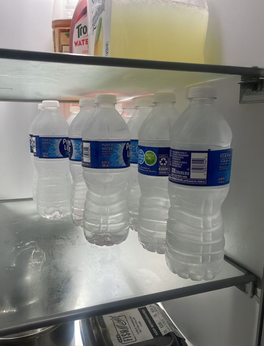 Five plastic water bottles are lined up on a refrigerator shelf, all partially filled and slightly squished. A carton and a jug of yellow liquid are visible on the shelf above.