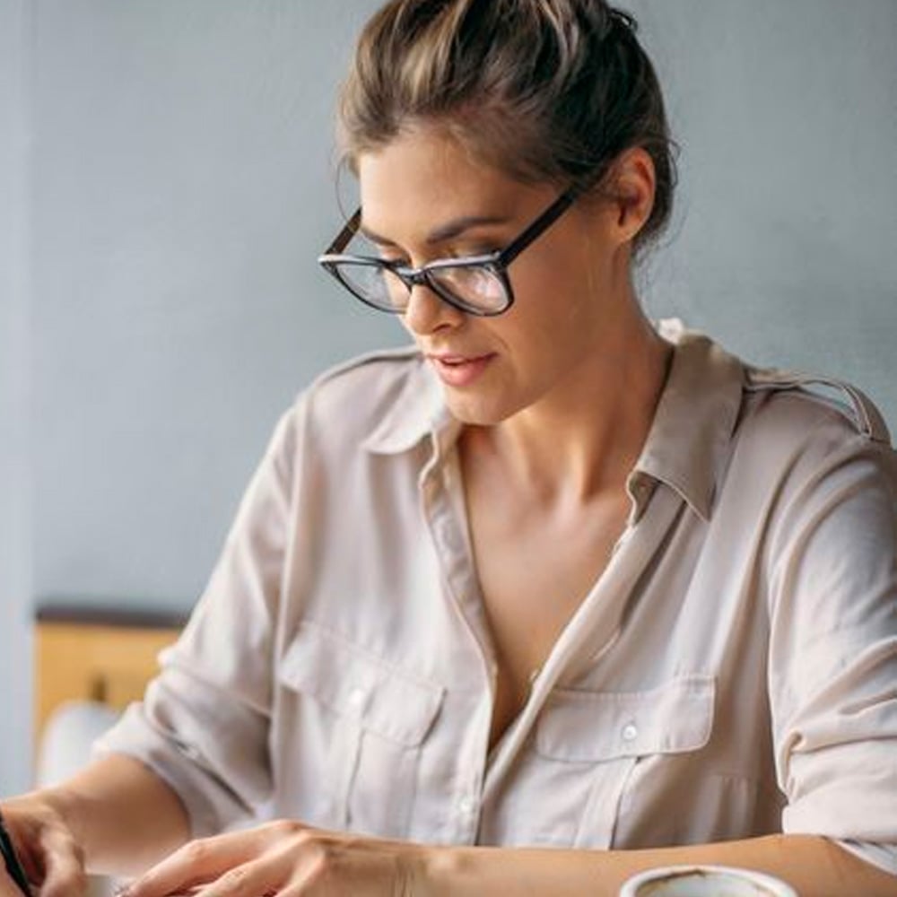 A woman with glasses and a loose, light-colored shirt is sitting at a table, focused on writing or reading something in front of her. She has her hair tied up and appears to be working or studying.