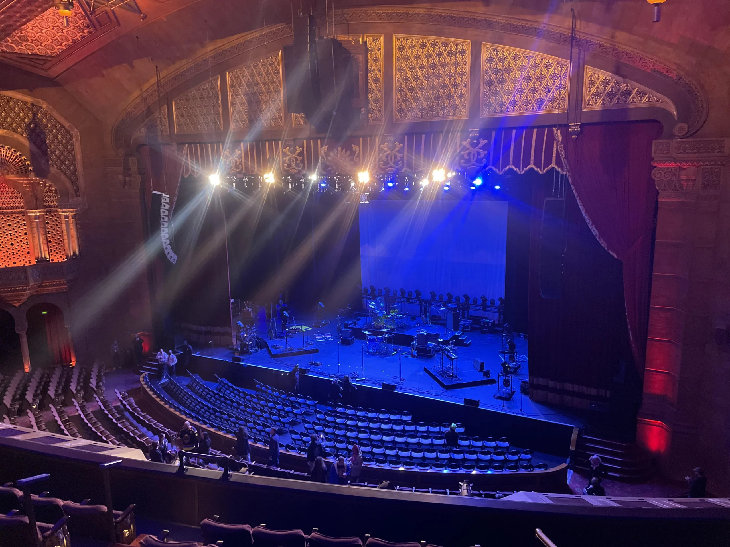 A theater stage lit with blue and yellow lights, featuring musical instruments set up for a performance. The ornate venue has red curtains, arched ceilings, and rows of seats, with a few people scattered throughout.