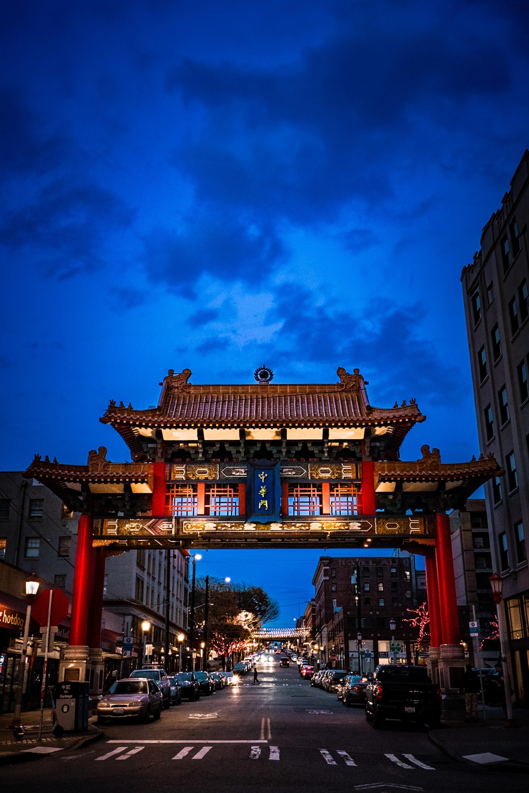 A traditional Chinese archway with red pillars and ornate decorations spans a city street at dusk, with cars parked along the road and buildings on either side under a deep blue sky.