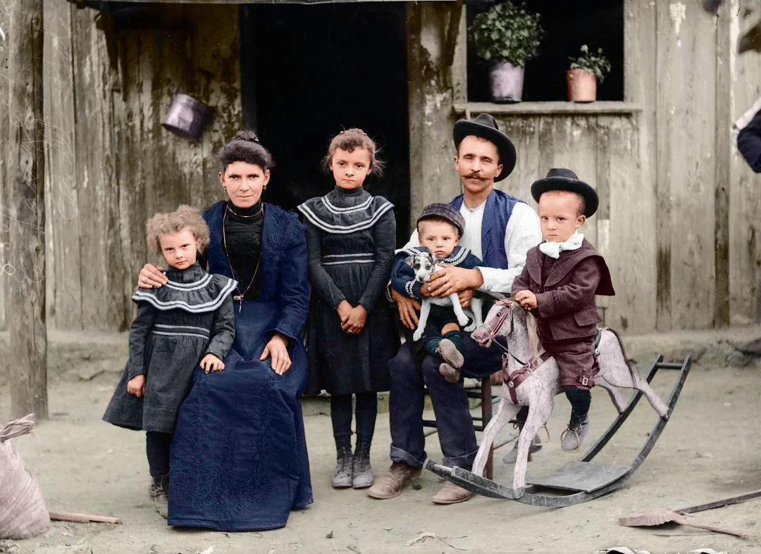 A vintage colorized photo of a family in front of a wooden house: a woman and three girls in dark dresses, a man holding a baby, and a boy on a wooden rocking horse. The scene appears early 20th century.