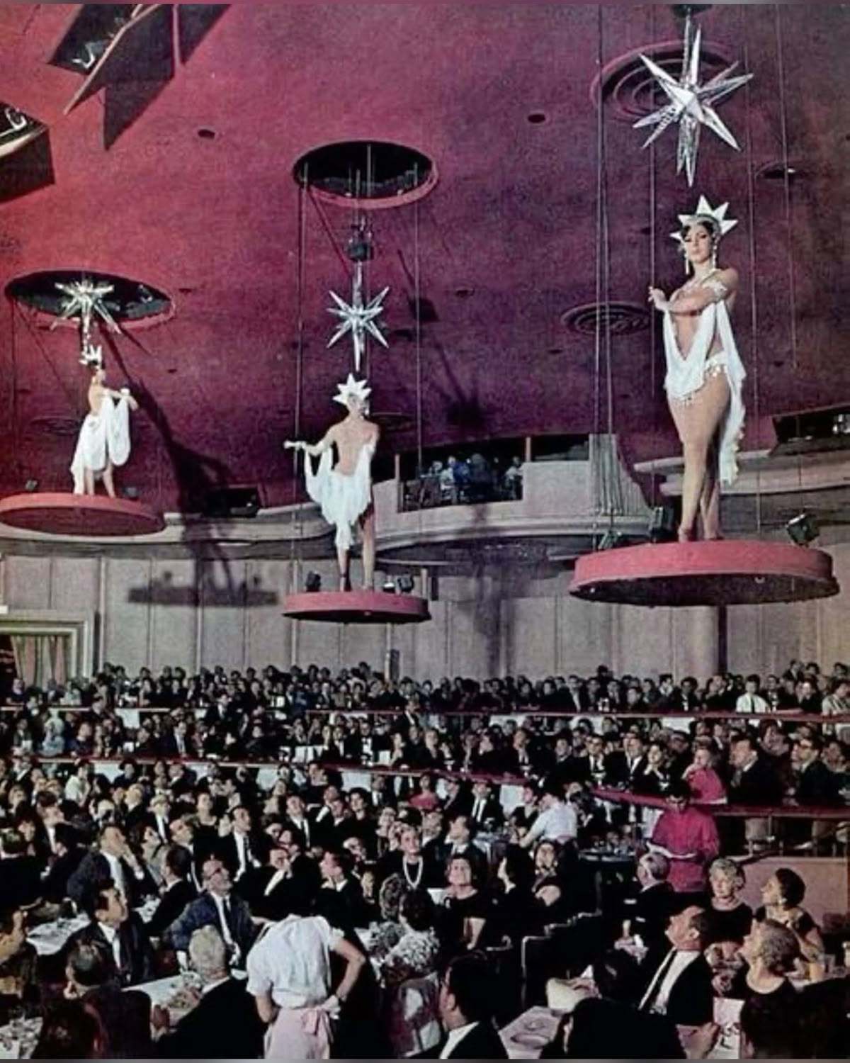 Showgirls in white costumes and star headpieces stand on large suspended platforms above a crowded, formal audience in a theater or nightclub with a red ceiling and hanging star decorations.