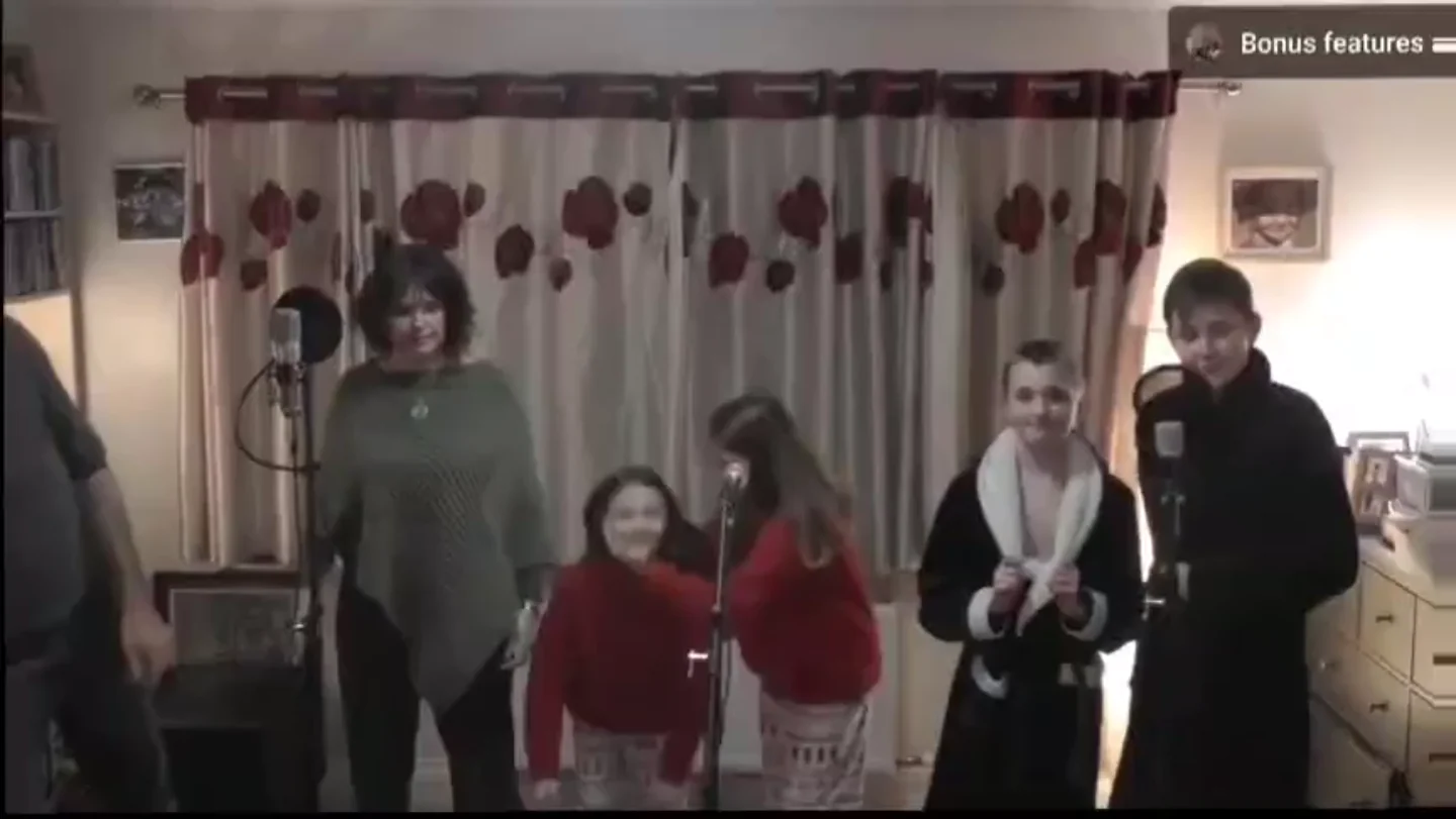 Six people stand and smile in a living room with floral curtains. Two children in red sweaters crouch near a microphone. Others stand behind or beside them, some wearing robes or sweaters. Microphones are visible in front of the group.