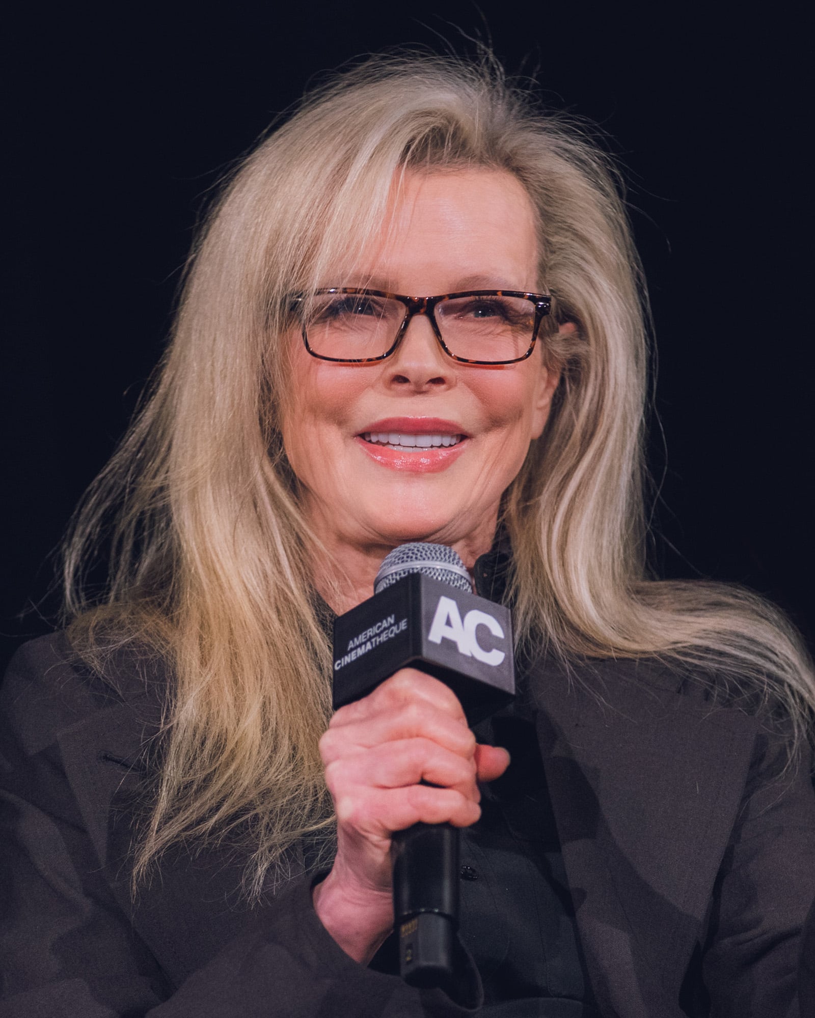 A woman with long blonde hair and glasses smiles while holding a microphone with an "AC" logo, wearing a black outfit and sitting against a dark background.