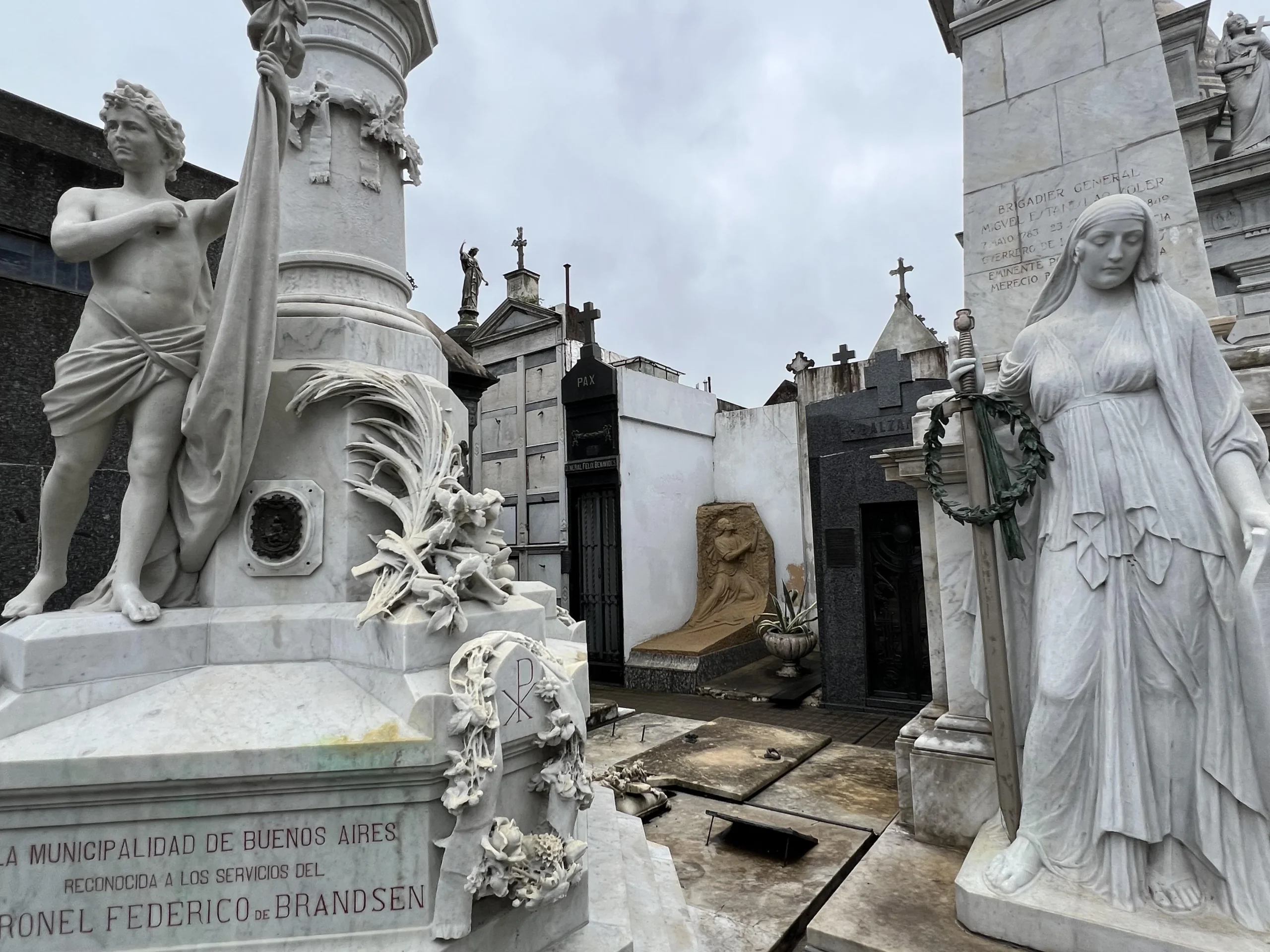 A cloudy day at a historic cemetery with ornate white marble statues and tombs, including a robed woman holding a wreath and a standing figure with drapery, surrounded by various mausoleums and crosses.