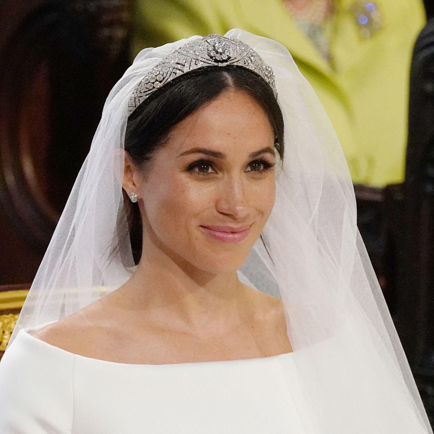 A bride wearing a white dress, diamond tiara, and veil smiles during a wedding ceremony. The background shows blurred figures, including a person in a bright yellow outfit.