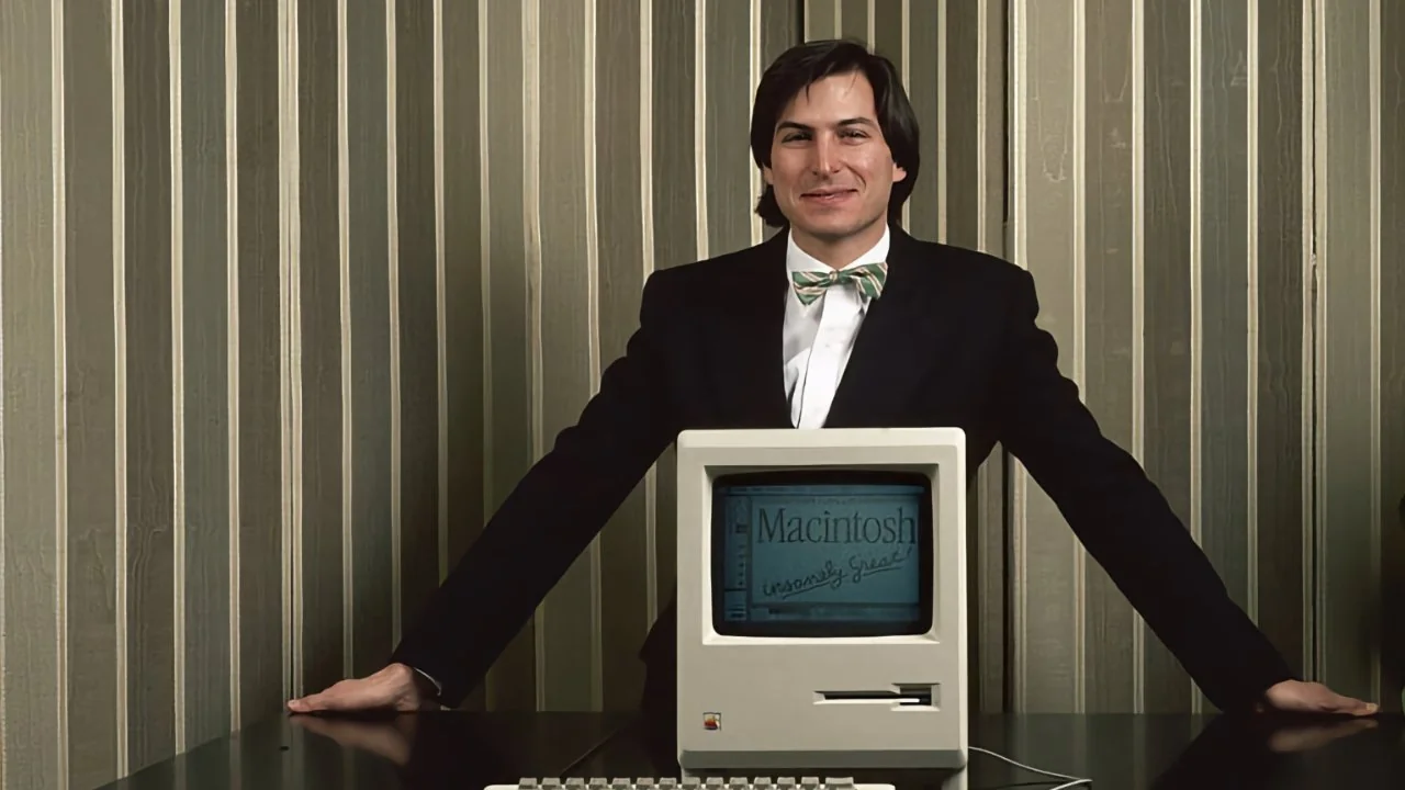 A man in a suit stands behind a vintage Macintosh computer with a keyboard and mouse on a table, in front of striped wallpaper. The computer screen displays "Macintosh.