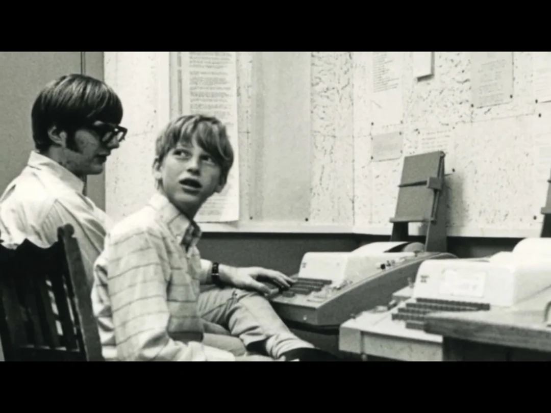 Two young people sit at vintage computer terminals in a black and white photo. One looks back over his shoulder while typing, and the other watches beside him. Papers and notes cover the wall in the background.