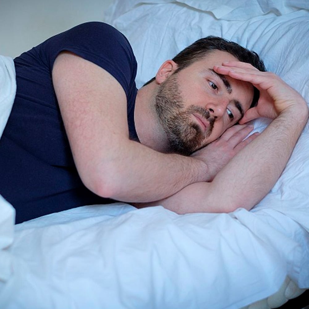 A man with a beard lies in bed on his side, resting his head on his hands and looking thoughtful or worried, with white sheets and a pillow around him.
