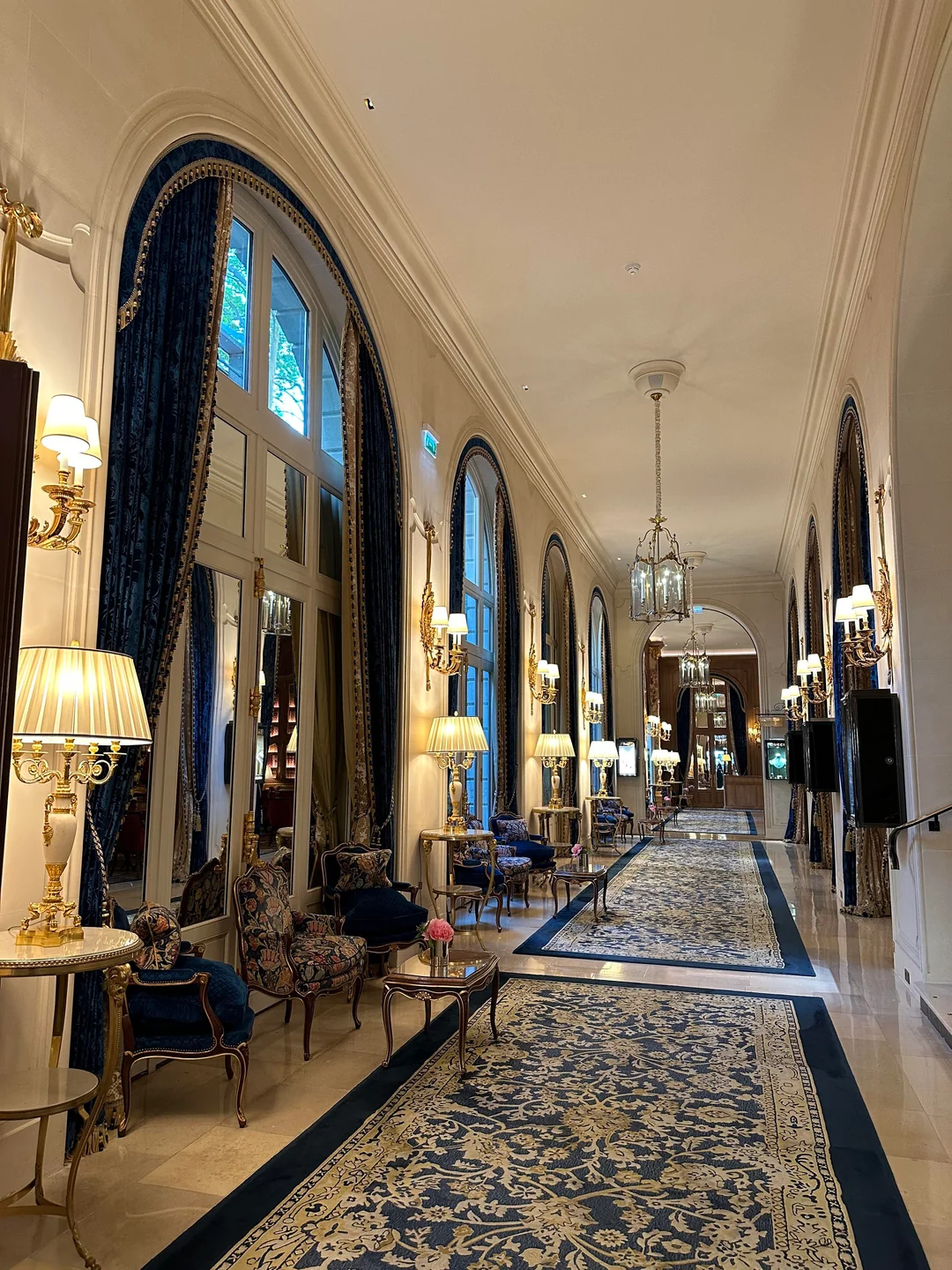 Elegant hotel hallway with large arched windows, blue velvet drapes, ornate chairs, gold lamps, chandeliers, and patterned rugs on a polished floor. Natural light streams in, creating a luxurious, classic atmosphere.