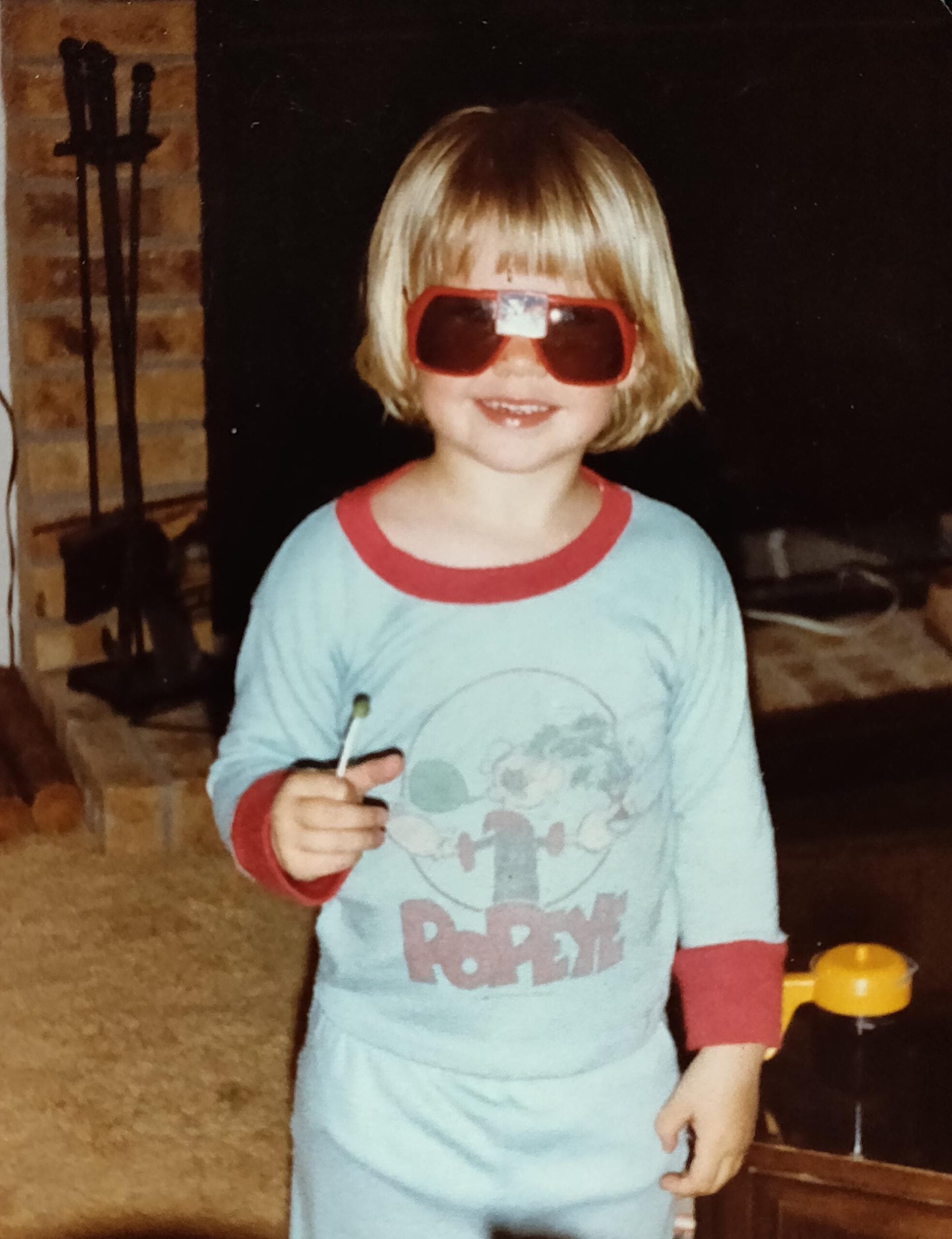 A young child wearing red sunglasses, light blue pajamas with a Popeye graphic, and holding a lollipop, smiles indoors near a brick fireplace and dark television.