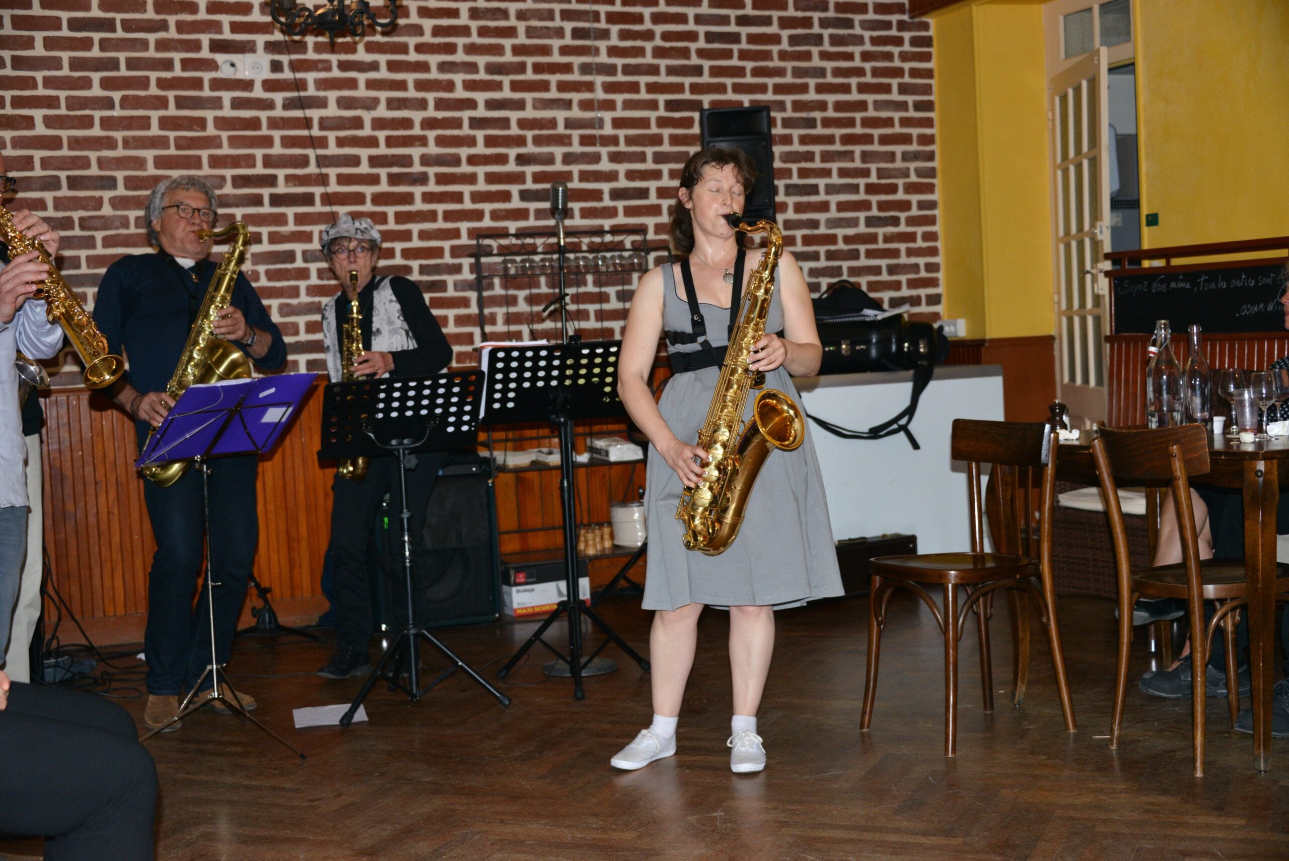 A small jazz band performs indoors; three musicians play saxophones. A woman in a gray dress stands in front, playing. Brick walls, tables, and chairs are visible in the cozy, warmly lit setting.