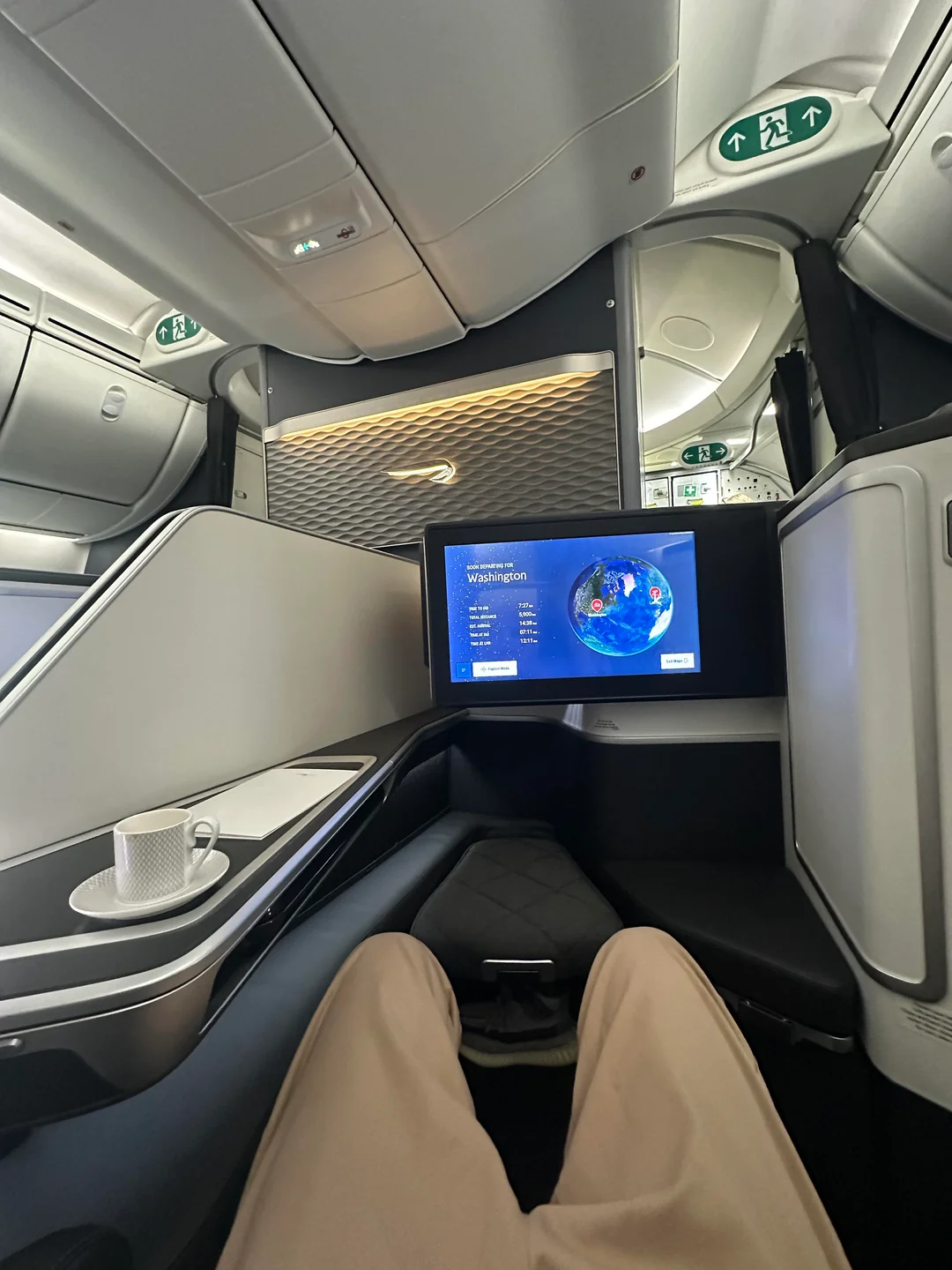 Person sitting in a first-class airplane seat with a cup on a side table, a large monitor displaying a flight map to Washington, and modern cabin interior visible.