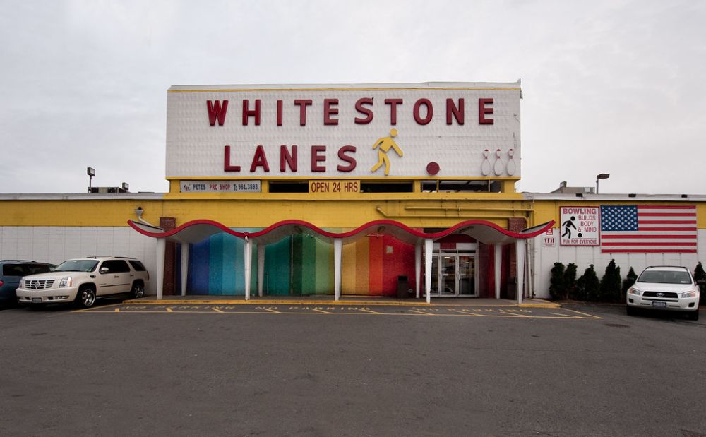 A large building with a sign reading "WHITESTONE LANES" above a colorful wavy awning; the entrance is below, with parking spaces and two cars in front. An American flag is displayed on the right.