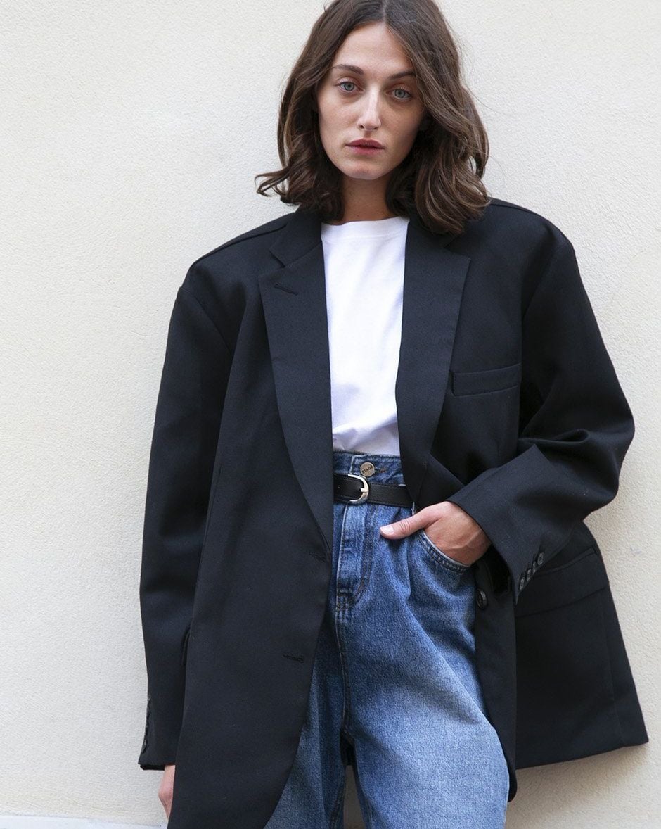 A person with shoulder-length brown hair wears an oversized black blazer over a white t-shirt, tucked into high-waisted blue jeans, standing against a plain light-colored wall.