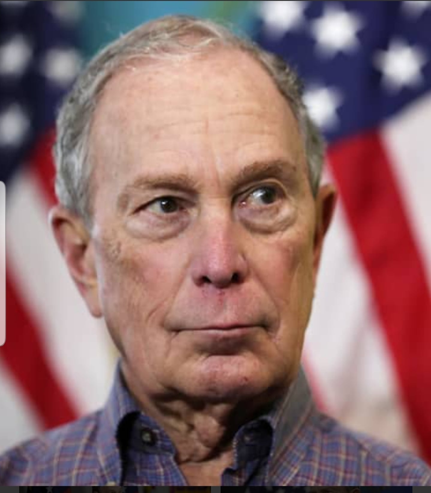 A man with short gray hair and a serious expression stands in front of blurred American flags. He is wearing a plaid collared shirt.