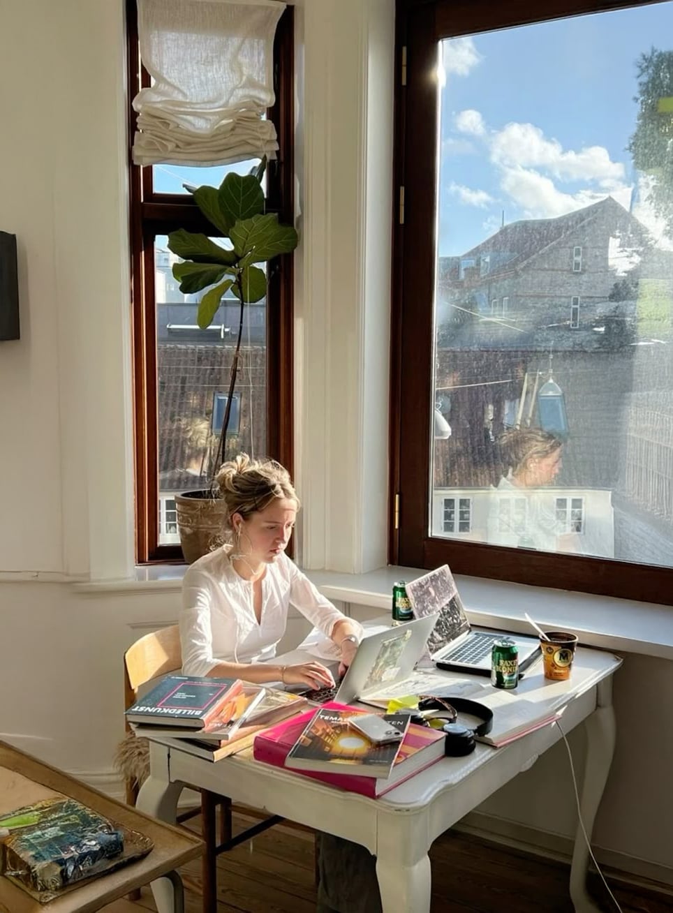 A woman sits at a white desk by large, sunny windows, working on a laptop surrounded by books, headphones, a coffee cup, and a plant. Sunlight streams into the cozy, bright room.